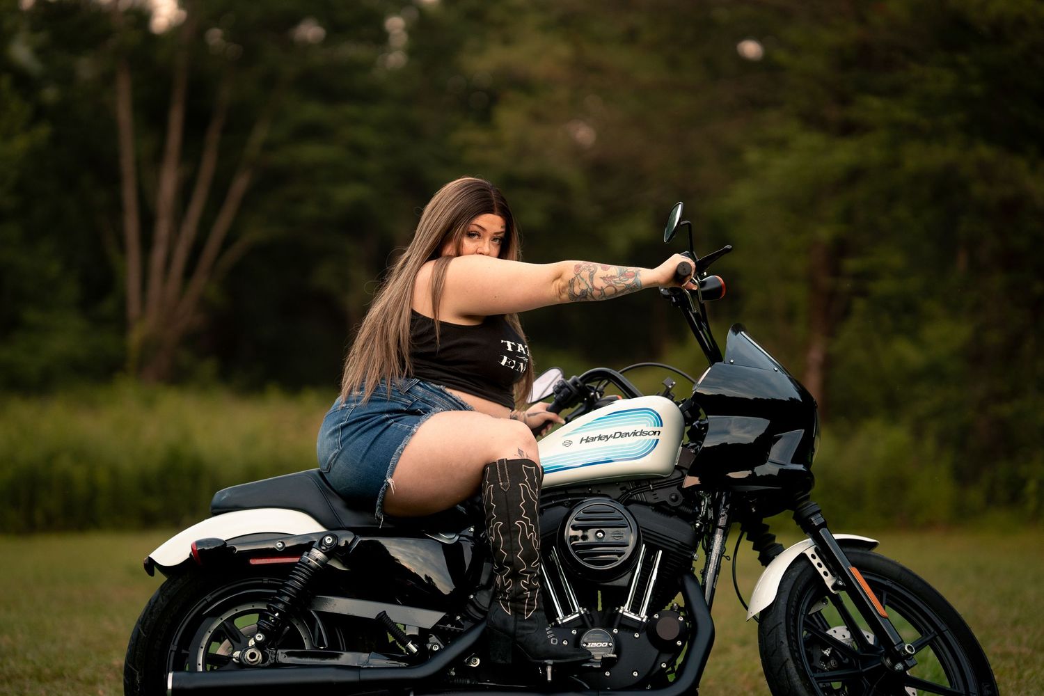 Woman with tattoos sitting on a Harley-Davidson motorcycle outdoors, wearing denim shorts and a black top.