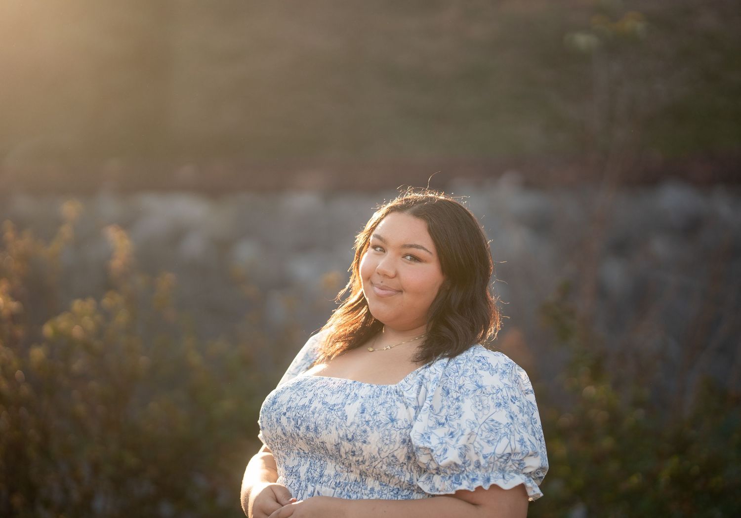Young woman in blue floral off-shoulder dress smiling in golden hour sunlight outdoors.