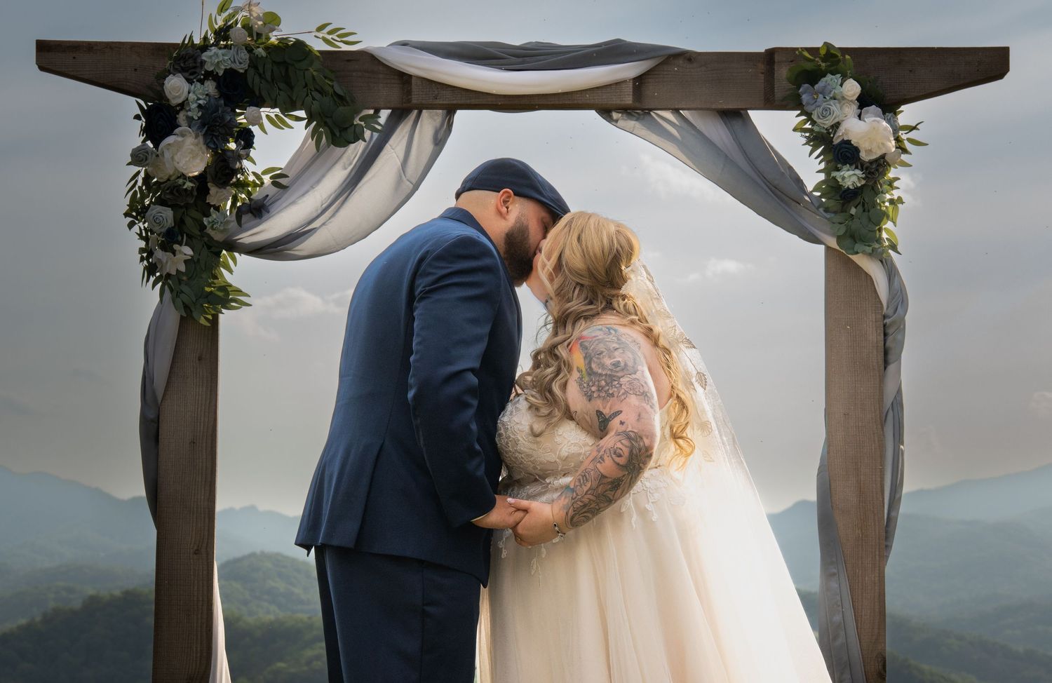 Bride and groom share a kiss under a rustic wooden arch with white florals and draping, overlooking misty mountain views.