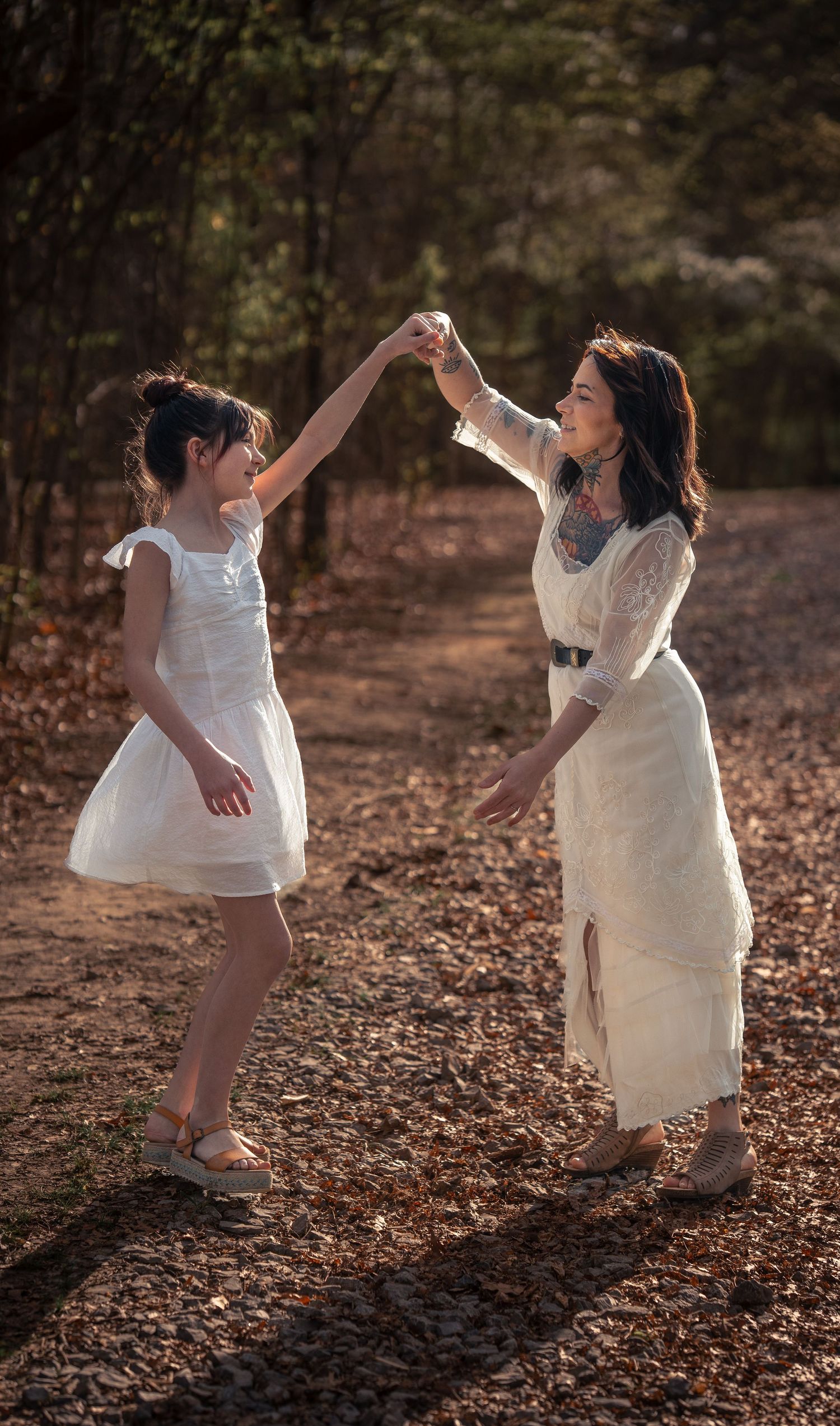 A mother and daughter in white dresses dance joyfully together on a sunlit forest path.