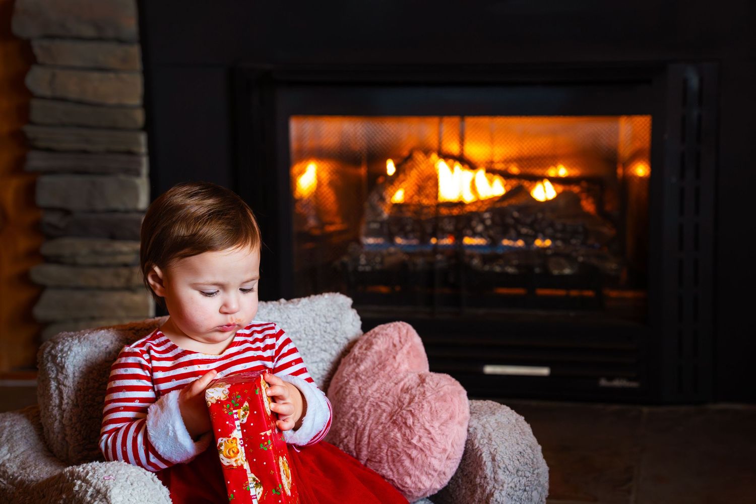 Baby girl in red tutu sitting by glowing fireplace holding Christmas gift.