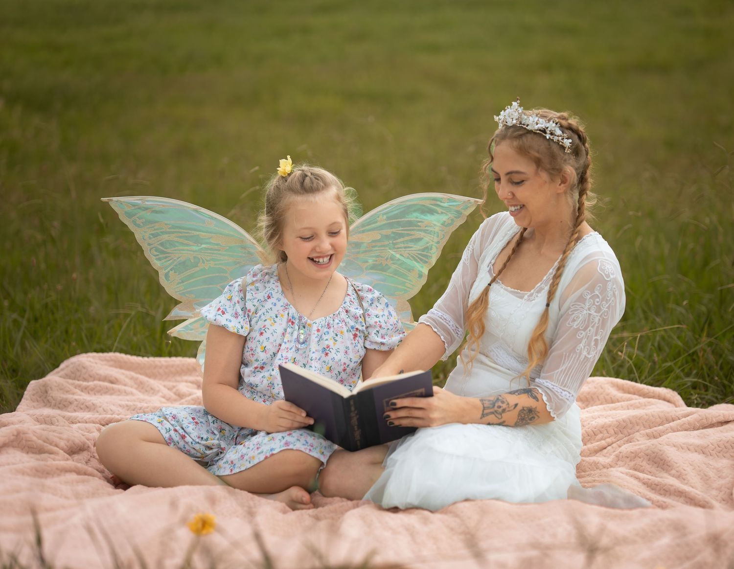 A woman and young girl dressed as fairies sit on a blanket in a field, smiling while reading a book together.