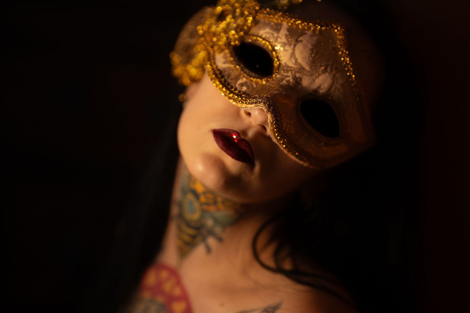 Tattooed woman in a gold masquerade mask with red lips, dramatic dark background portrait.