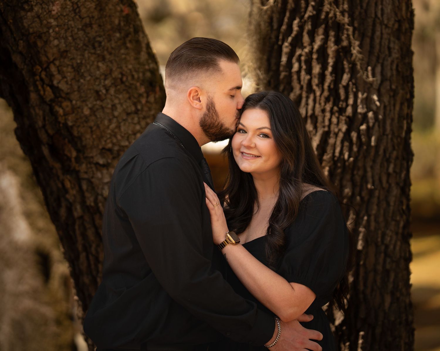 A romantic couple in black embracing and kissing near a tree in golden autumn light.