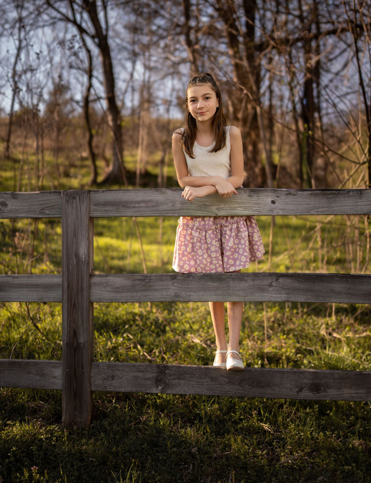 Young girl in white top and floral skirt poses by rustic wooden fence outdoors.