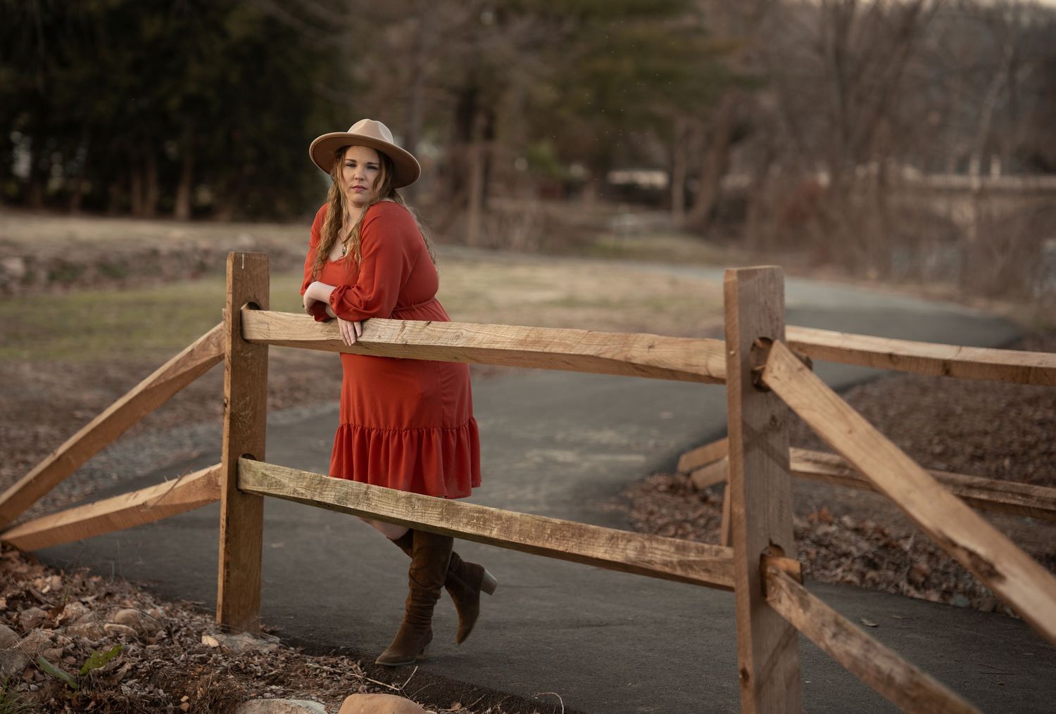 Woman in red dress and hat poses by rustic wooden fence near a country road.