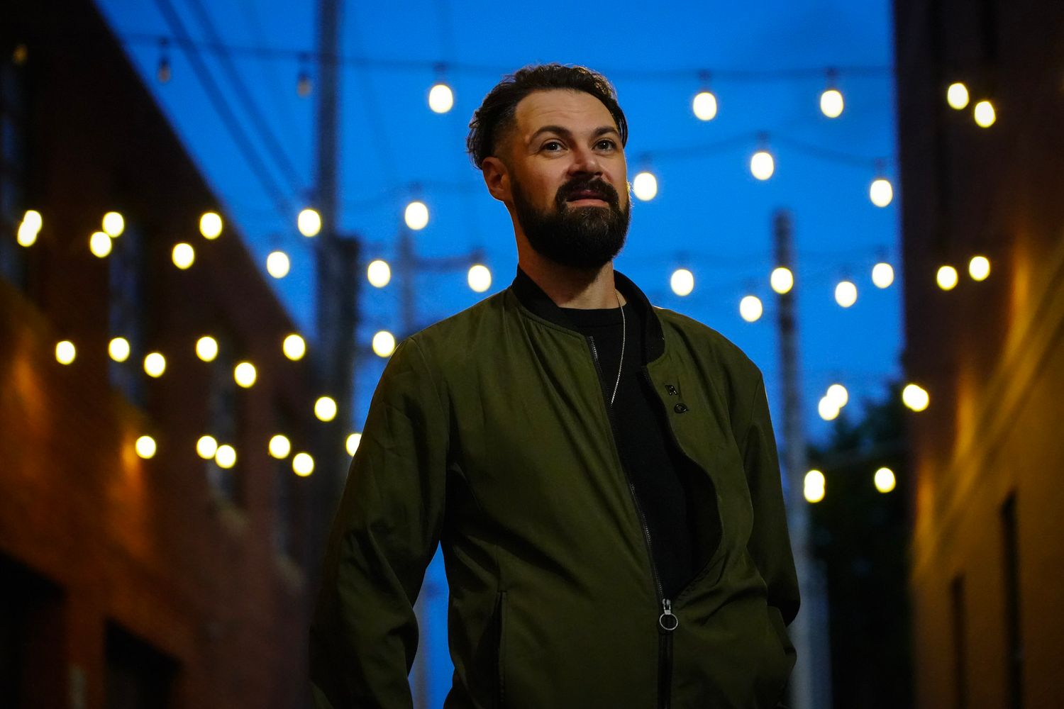 Bearded man in olive green bomber jacket standing in alley with string lights at dusk.