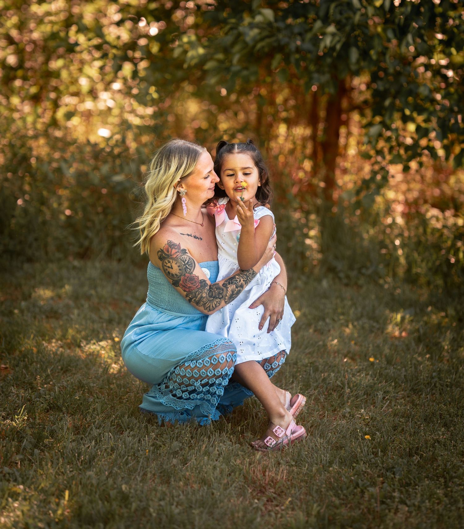 A tattooed blonde woman in a blue dress crouches outdoors, lovingly holding a young girl in a white dress.