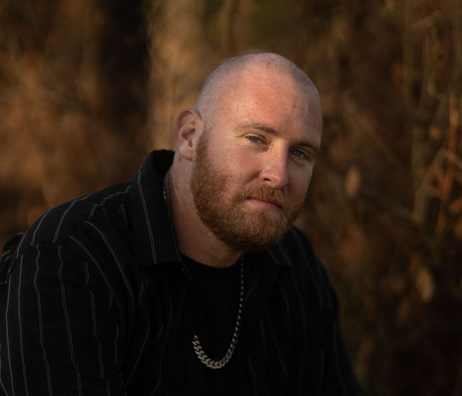 Bald man with red beard wearing striped shirt and silver chain necklace posing outdoors.
