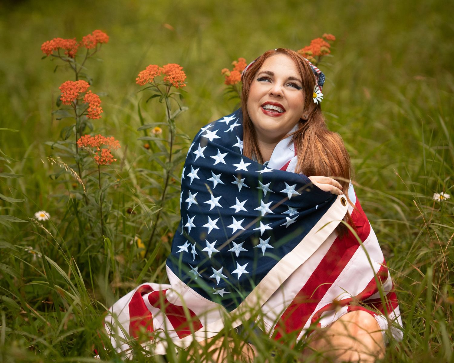 Smiling redhead woman wrapped in American flag sitting in green field with orange wildflowers.