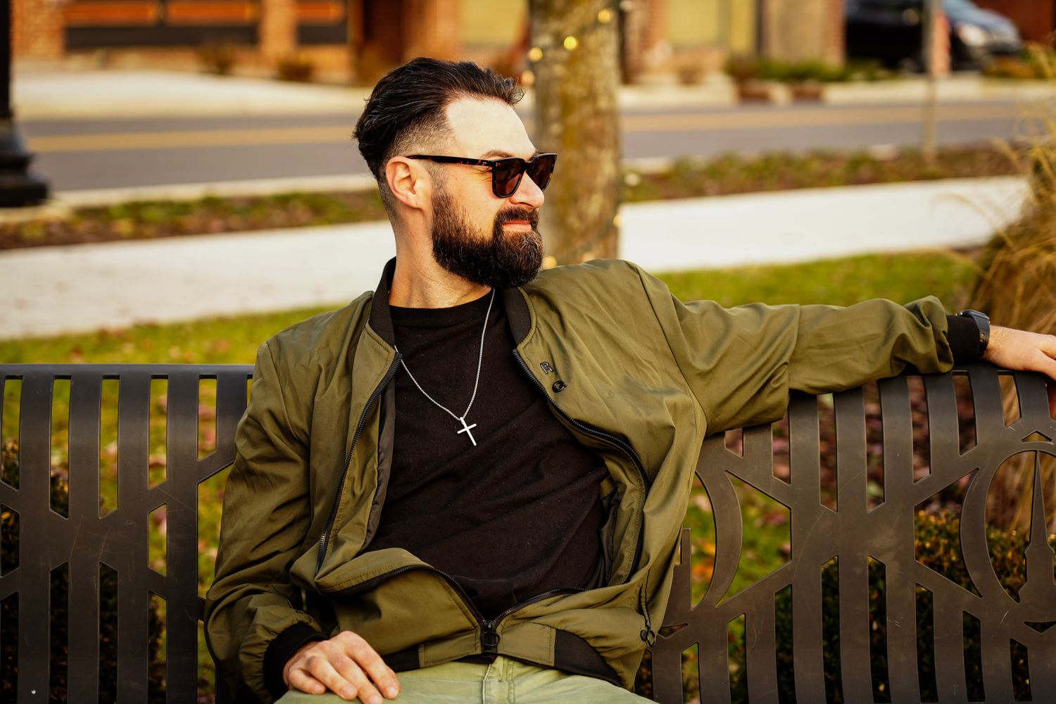 Bearded man in olive jacket and sunglasses relaxing on park bench wearing cross necklace.