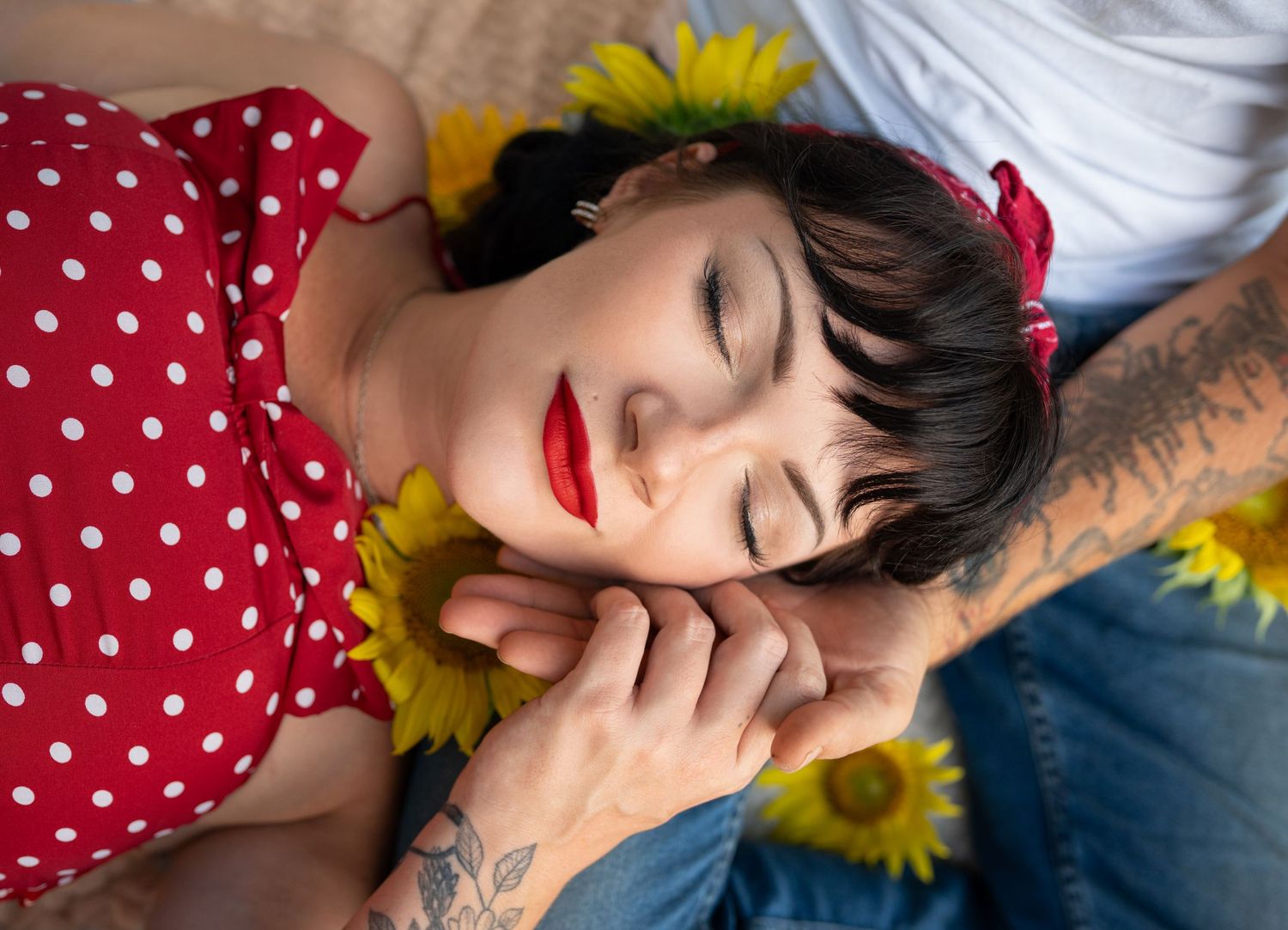 Woman with red lips and polka dot top resting among sunflowers, with tattooed arms visible nearby.