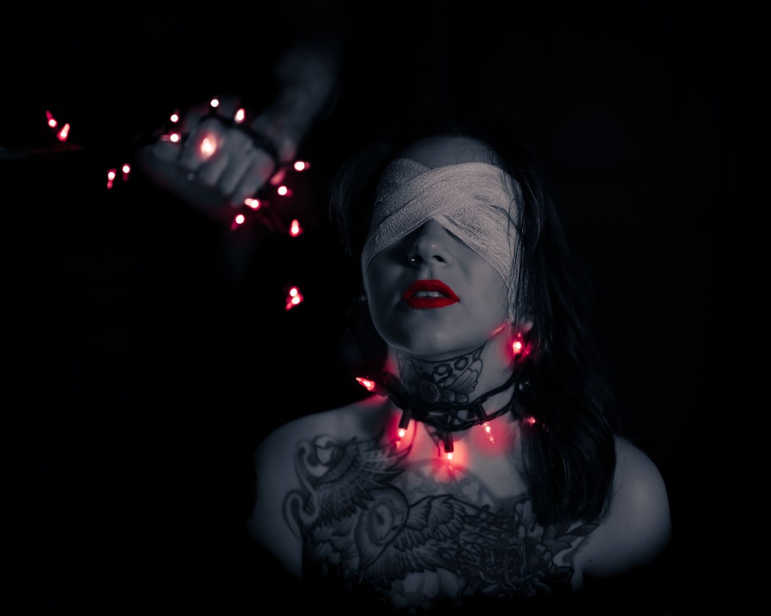 Tattooed woman with bandaged eyes and red string lights around her neck in a dark, moody setting.