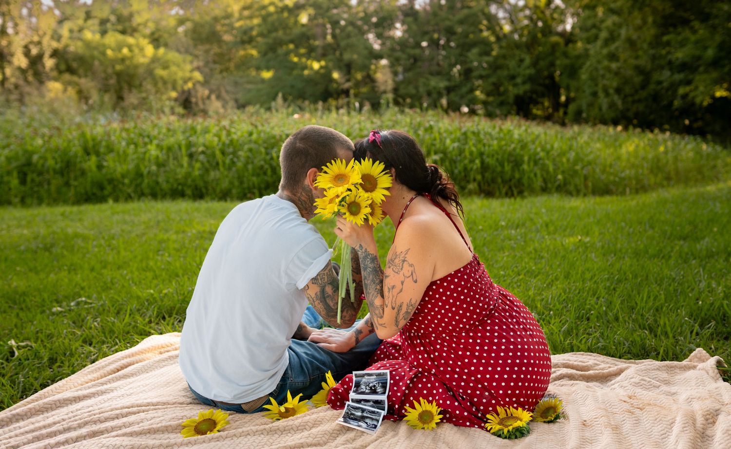 Couple sitting on blanket in park with sunflowers and ultrasound photo for maternity announcement.