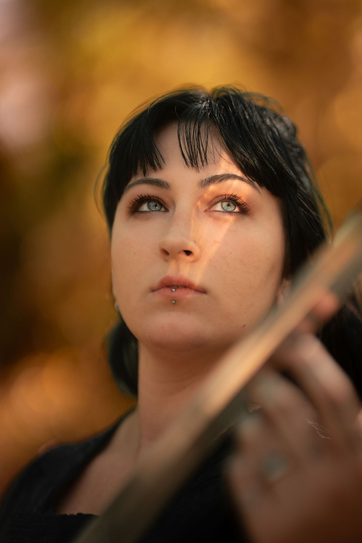 Dark-haired woman with blue eyes holding a sword looking upward in autumn light.