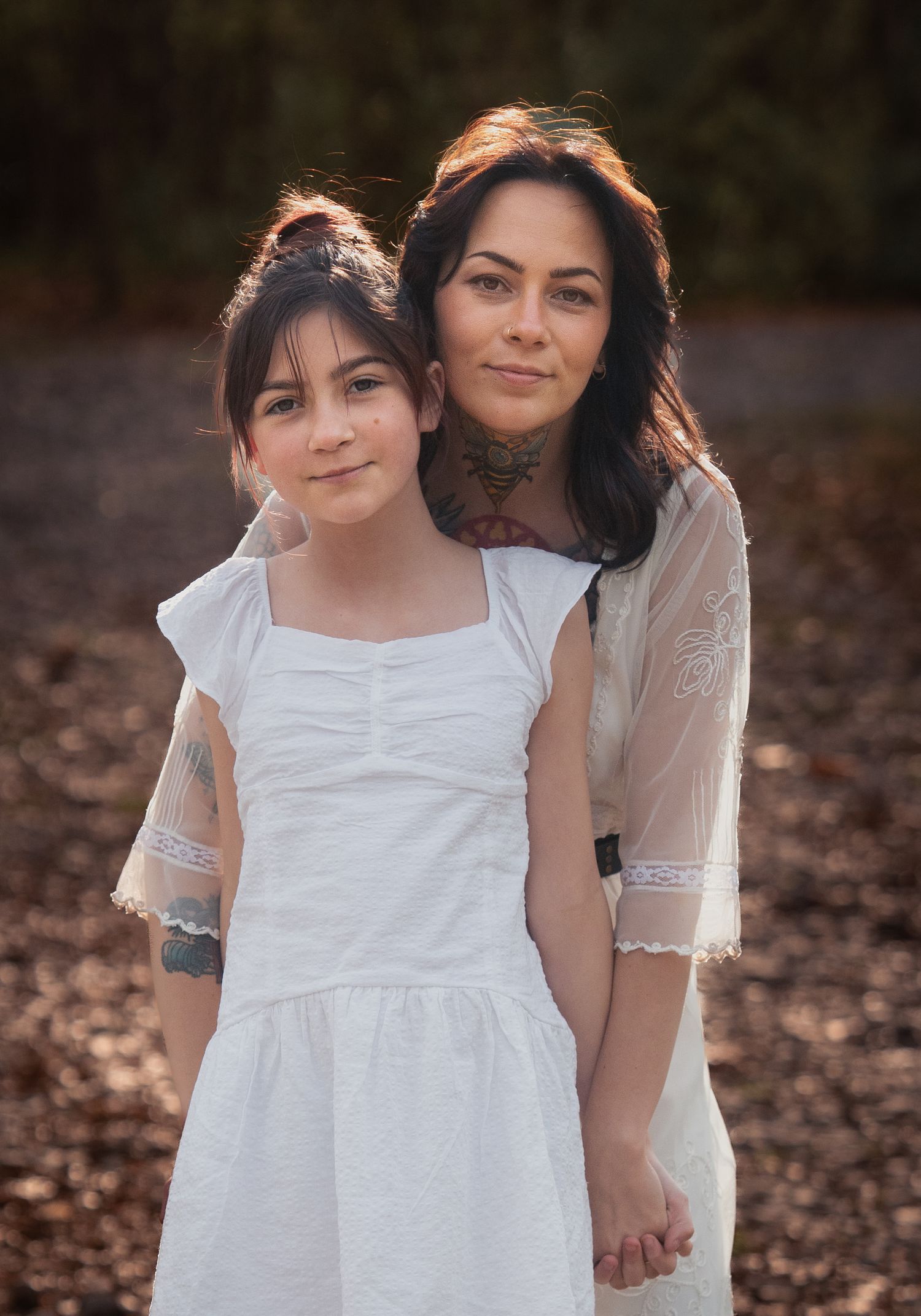 A mother and daughter in matching white dresses pose together outdoors among autumn leaves.