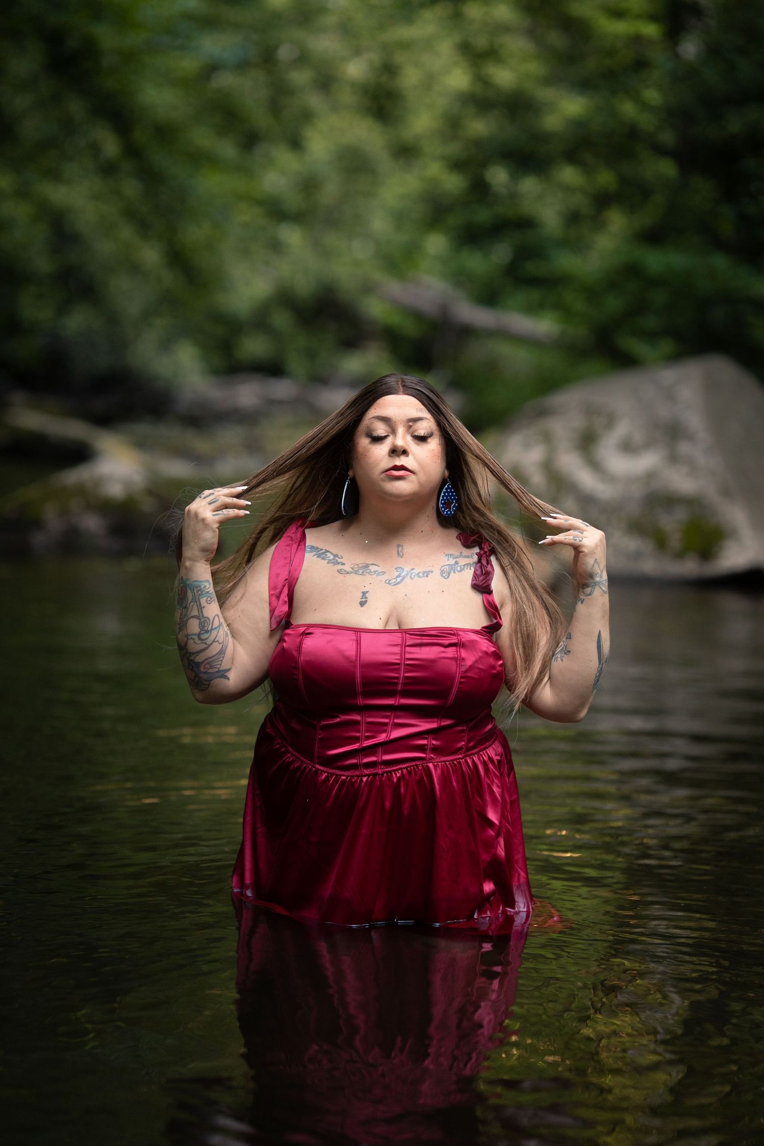 Woman in red satin dress standing in a forest river, with tattoos and blue earrings, posing with eyes closed.