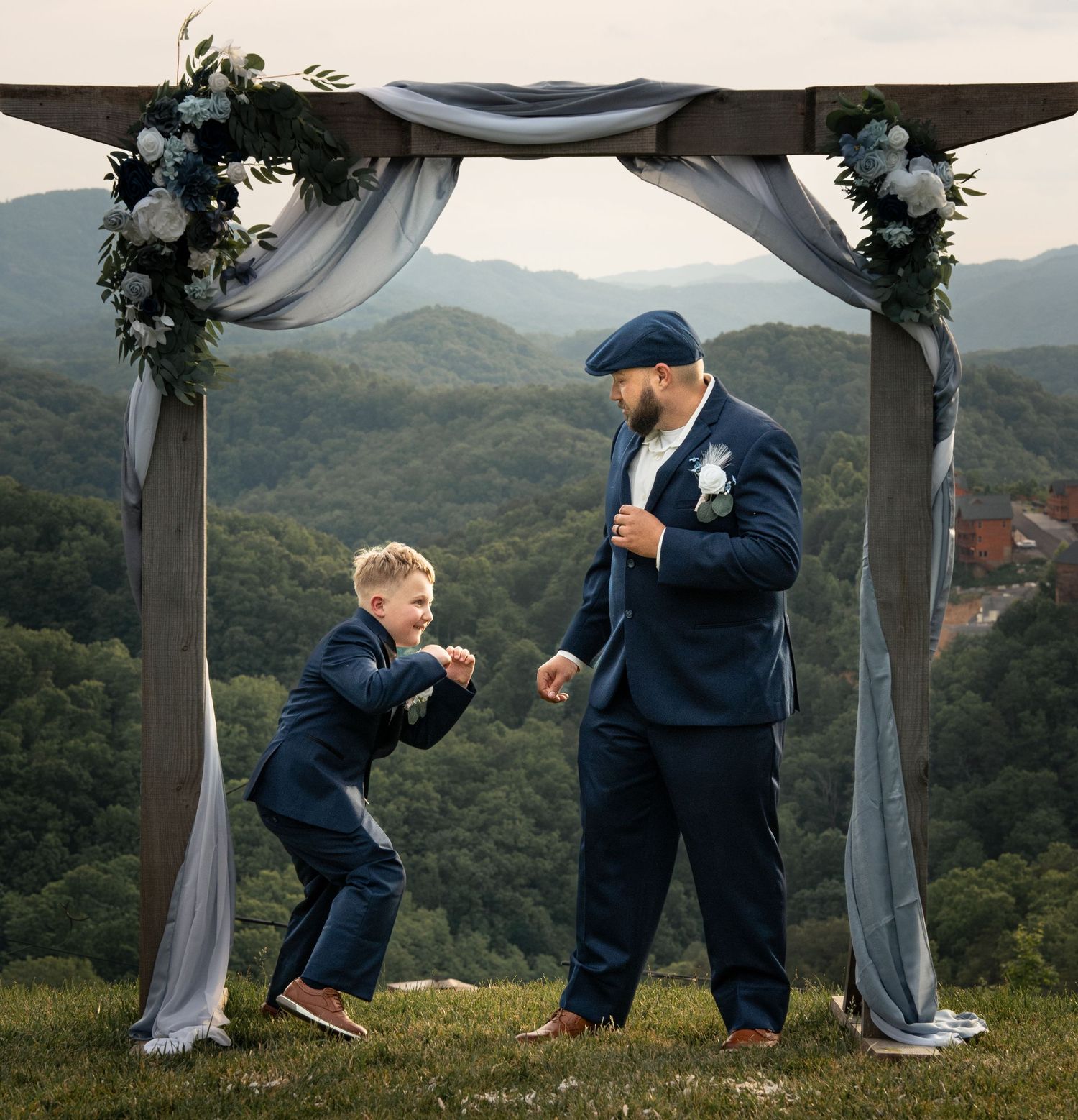 A man and young boy in matching navy suits share a playful moment under a floral wedding arch with mountain views.
