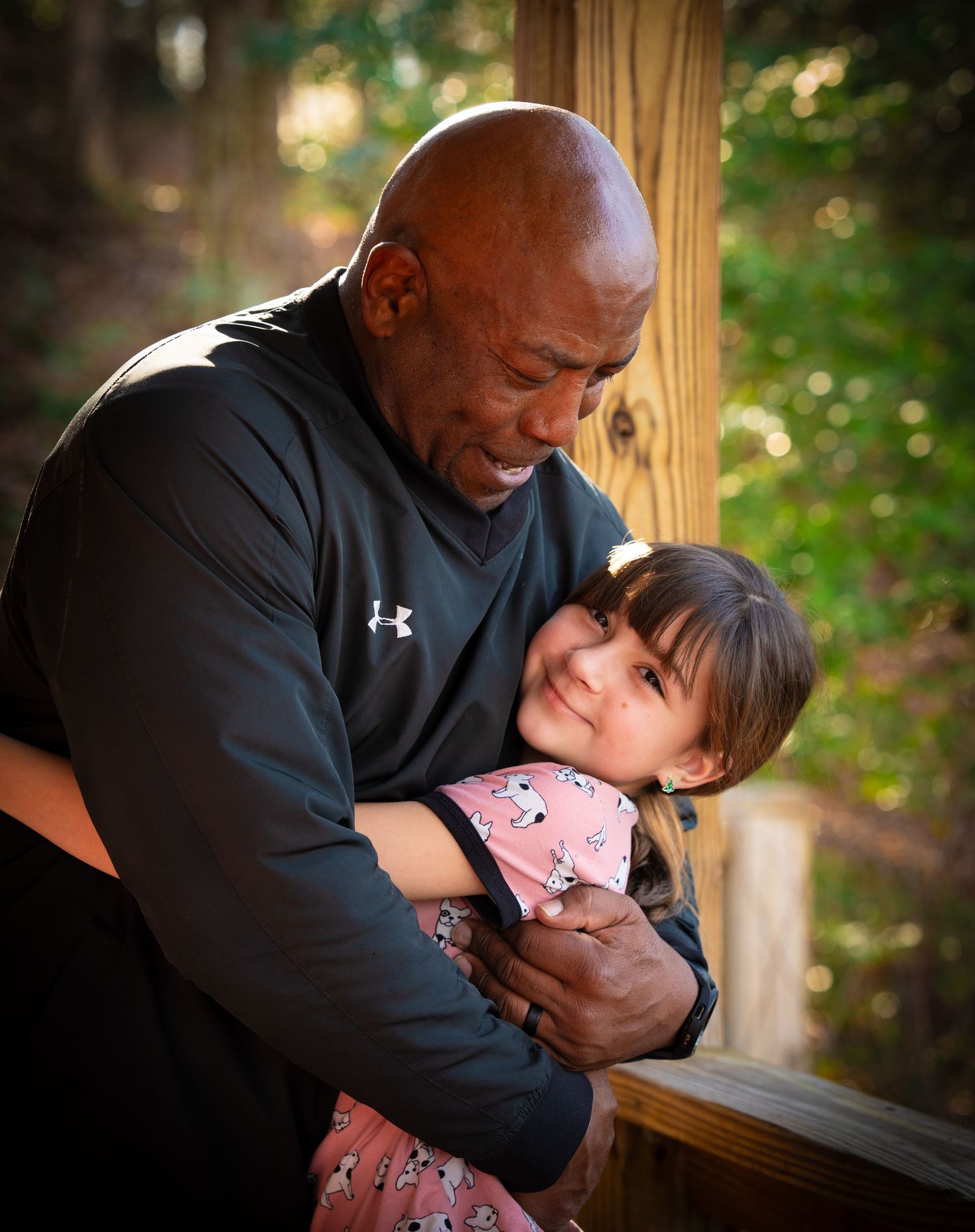 Bald man in black shirt hugging smiling young girl on wooden deck.