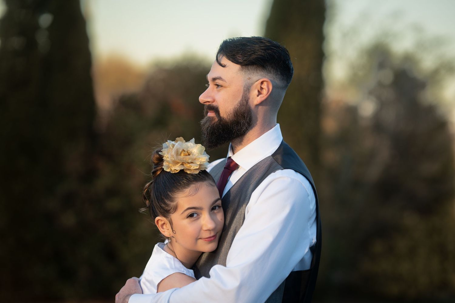 Smiling bearded man in vest hugs young girl with floral hair accessory outdoors.