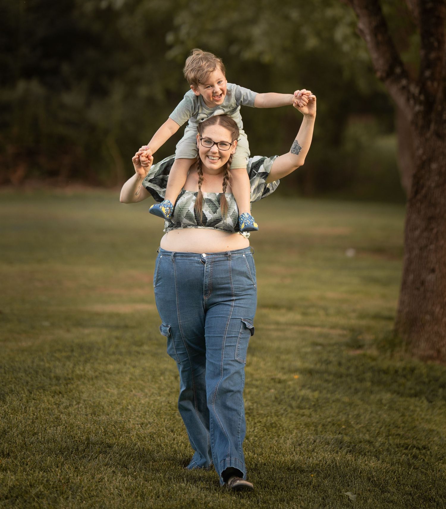 Woman carrying smiling boy on her shoulders while walking through a lush green park on a sunny day.