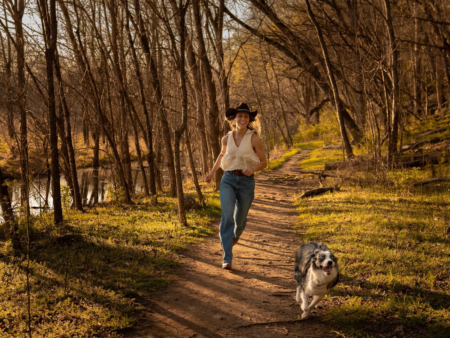 Woman in cowboy hat running with Australian Shepherd dog along sunny wooded trail.