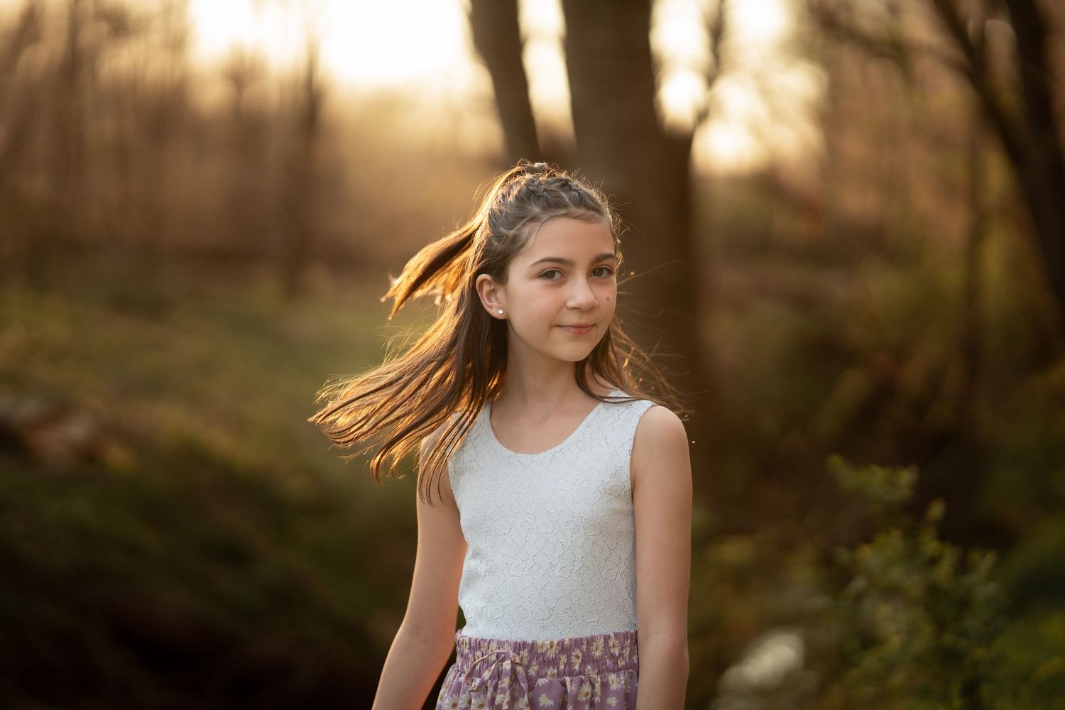 Young girl in white top with flowing hair poses in golden sunset forest light.