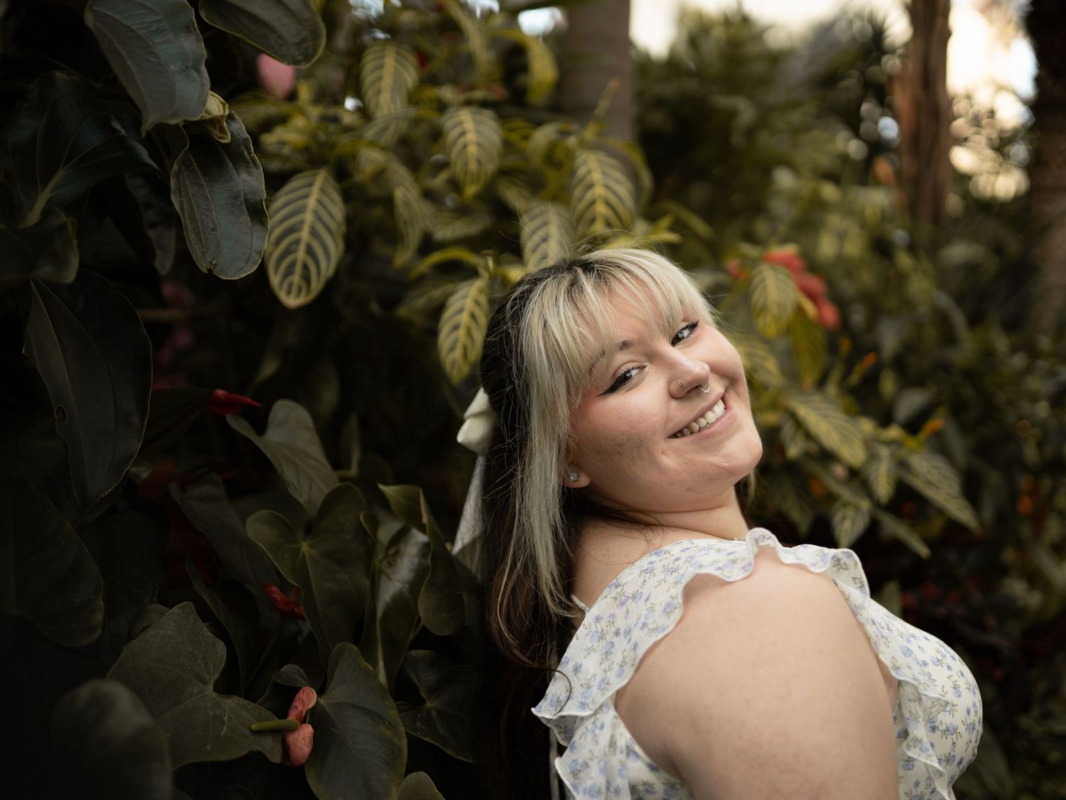Smiling woman in white ruffled dress peeking through lush tropical green foliage.