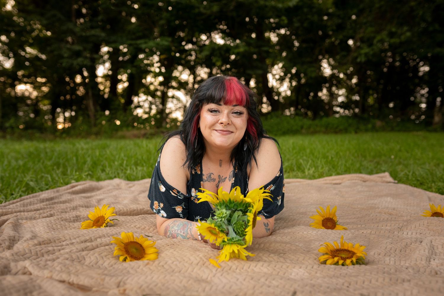 Woman with red-highlighted hair lying on a blanket outdoors, smiling with sunflowers around her in a green park.