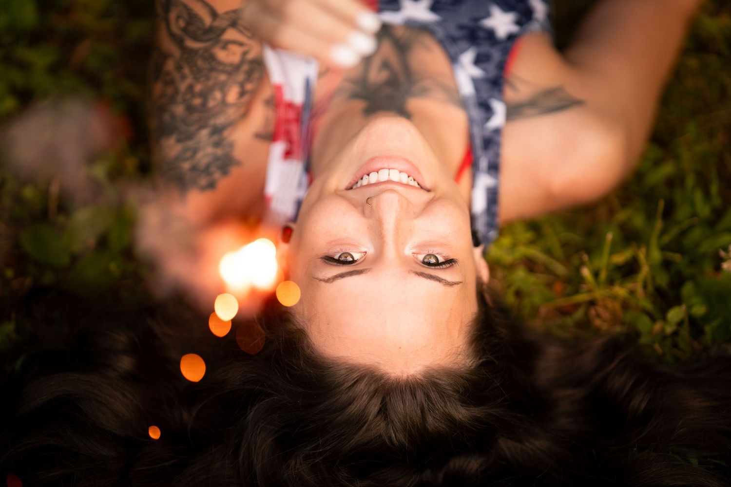 Tattooed woman lying on grass smiling upside down near sparkling bokeh lights outdoors.