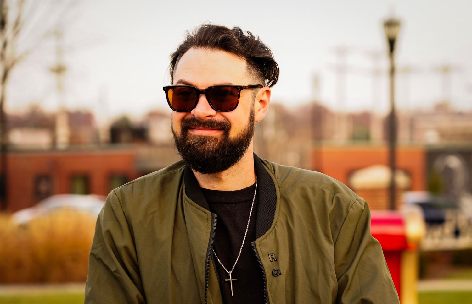 Smiling bearded man wearing sunglasses and olive jacket outdoors in urban setting.