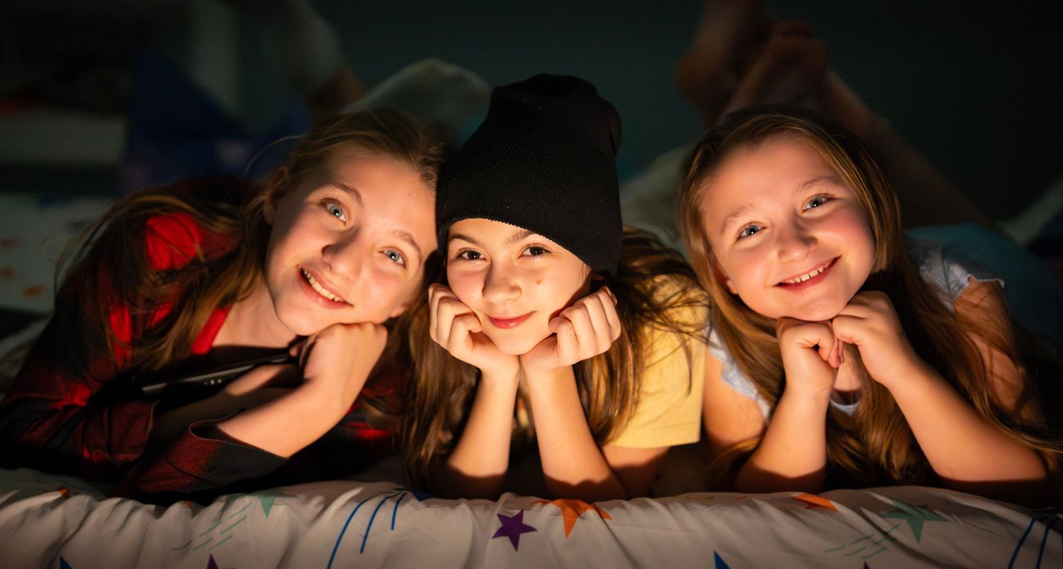 Three cheerful young girls smiling together while lying on a bed at a sleepover.