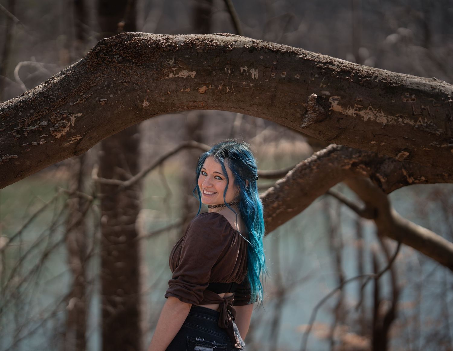 Smiling woman with blue hair looks back over shoulder near curved tree branch.