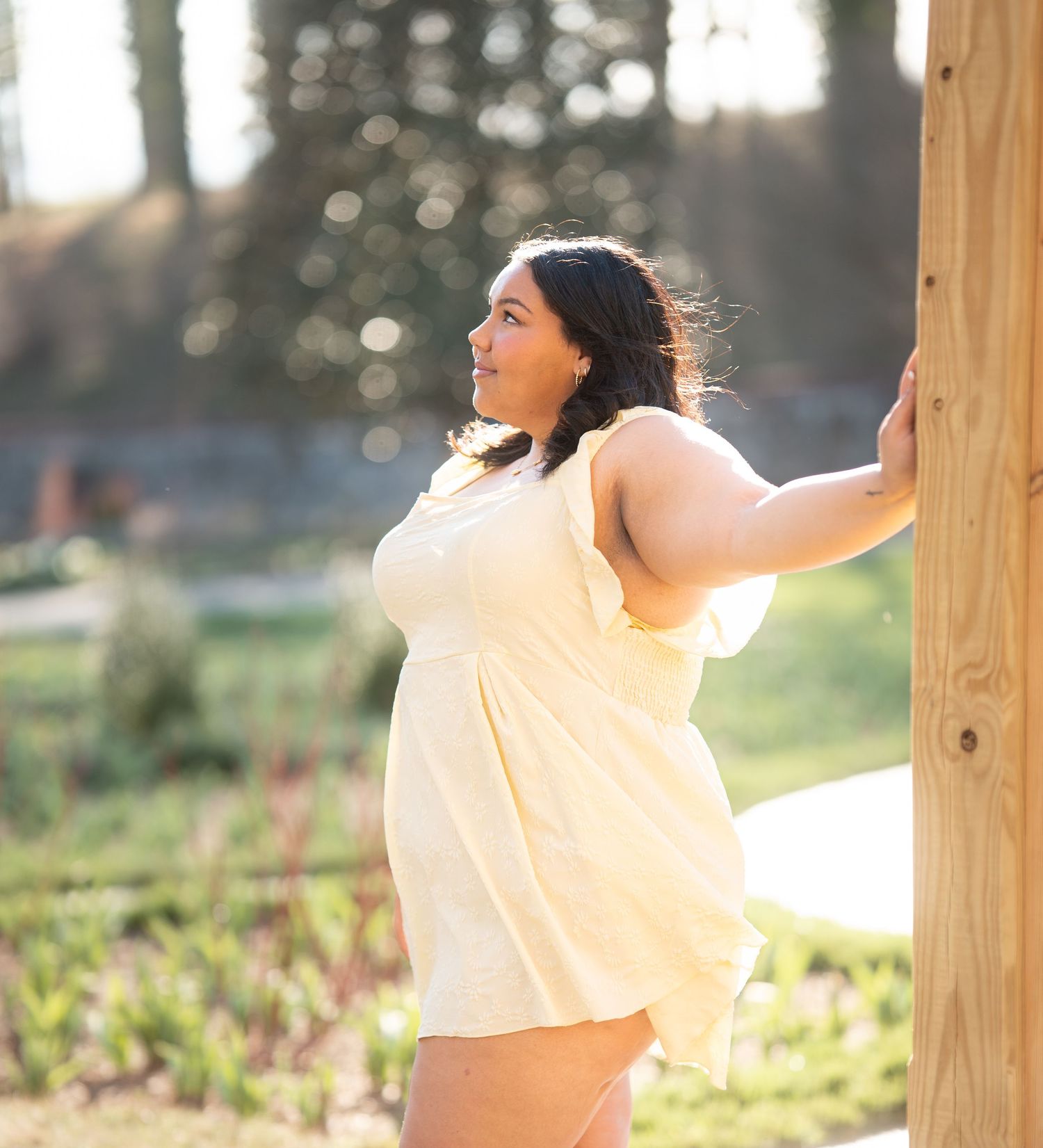 Plus size woman in yellow sundress leaning against wooden post in sunny outdoor garden setting.