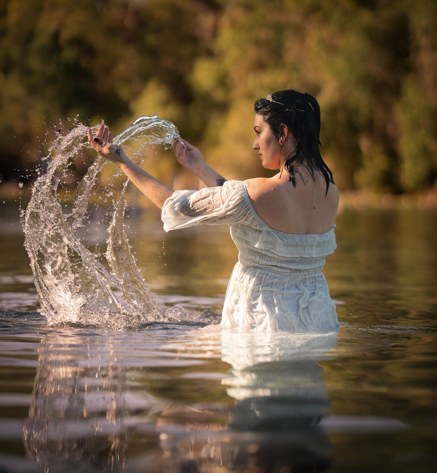 Woman in white off-shoulder dress splashing water while standing in a sunlit river.