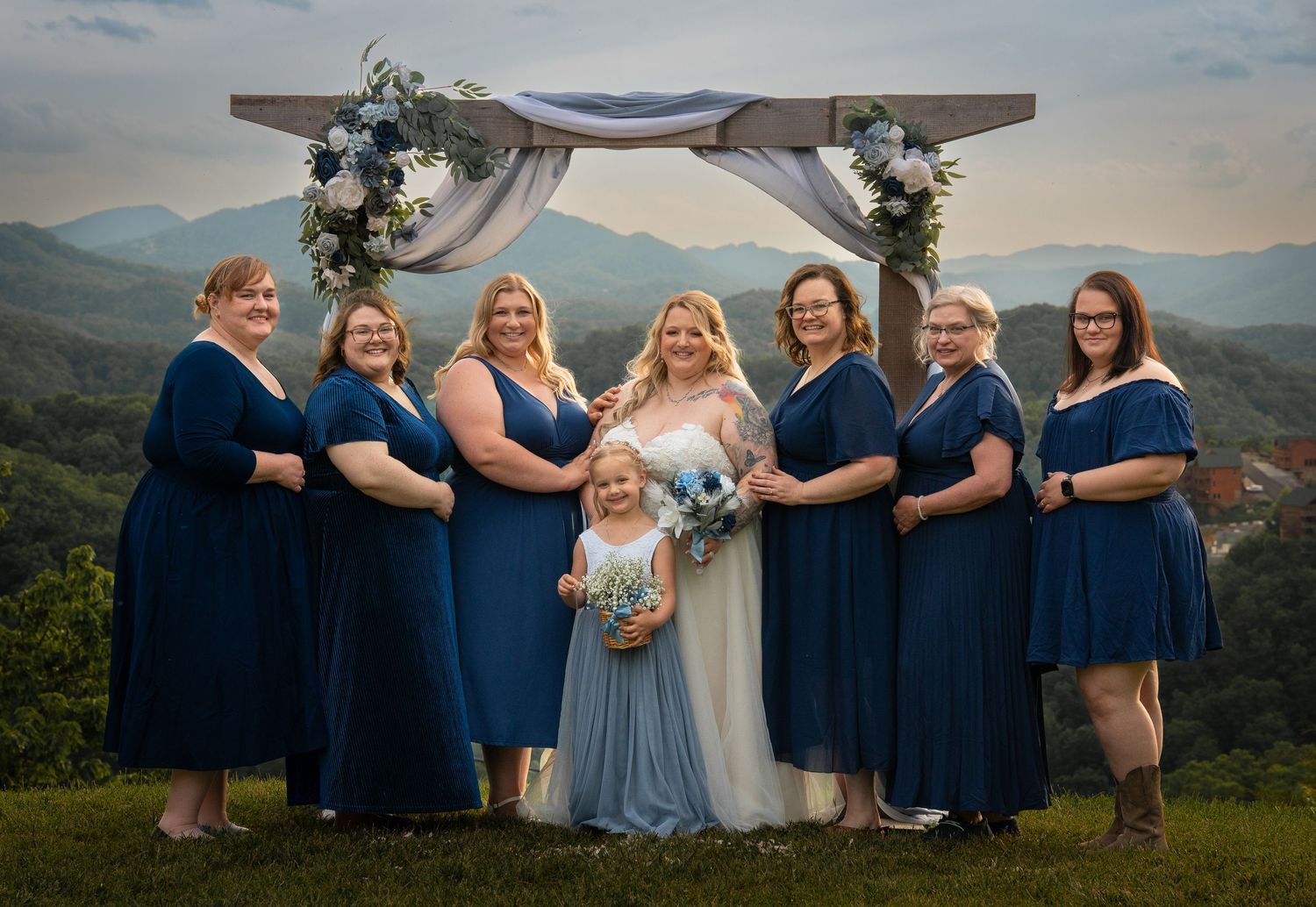 Bride in blue tulle gown with bridesmaids in navy dresses posing under floral arch with mountain views.