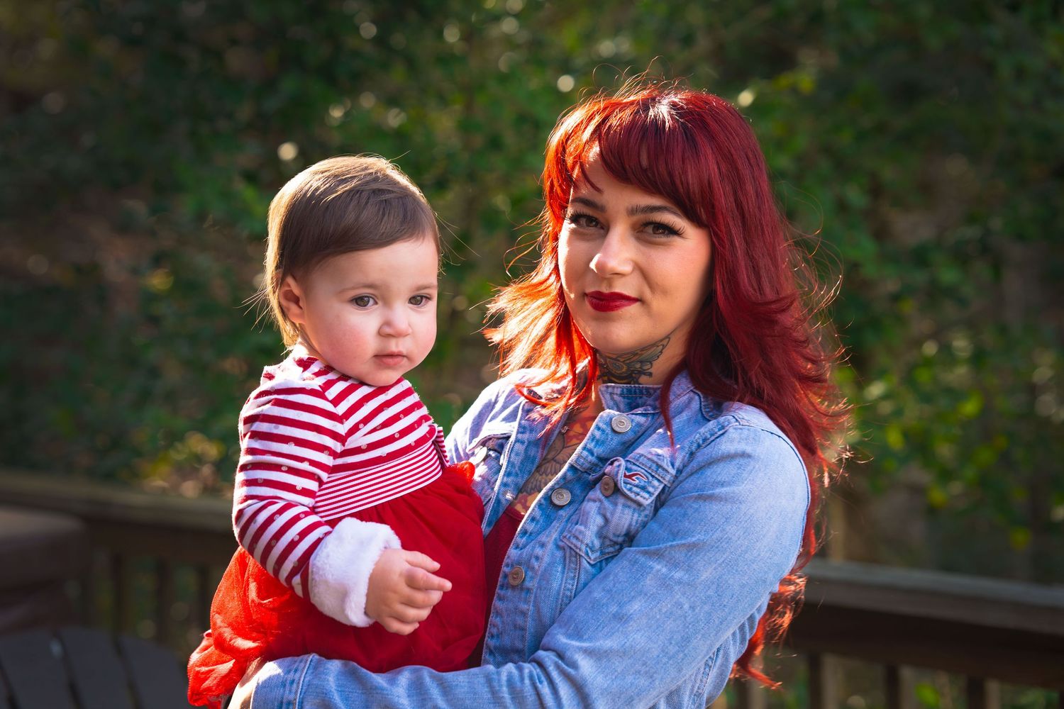 Red-haired mother in denim jacket holding baby in red outfit outdoors.