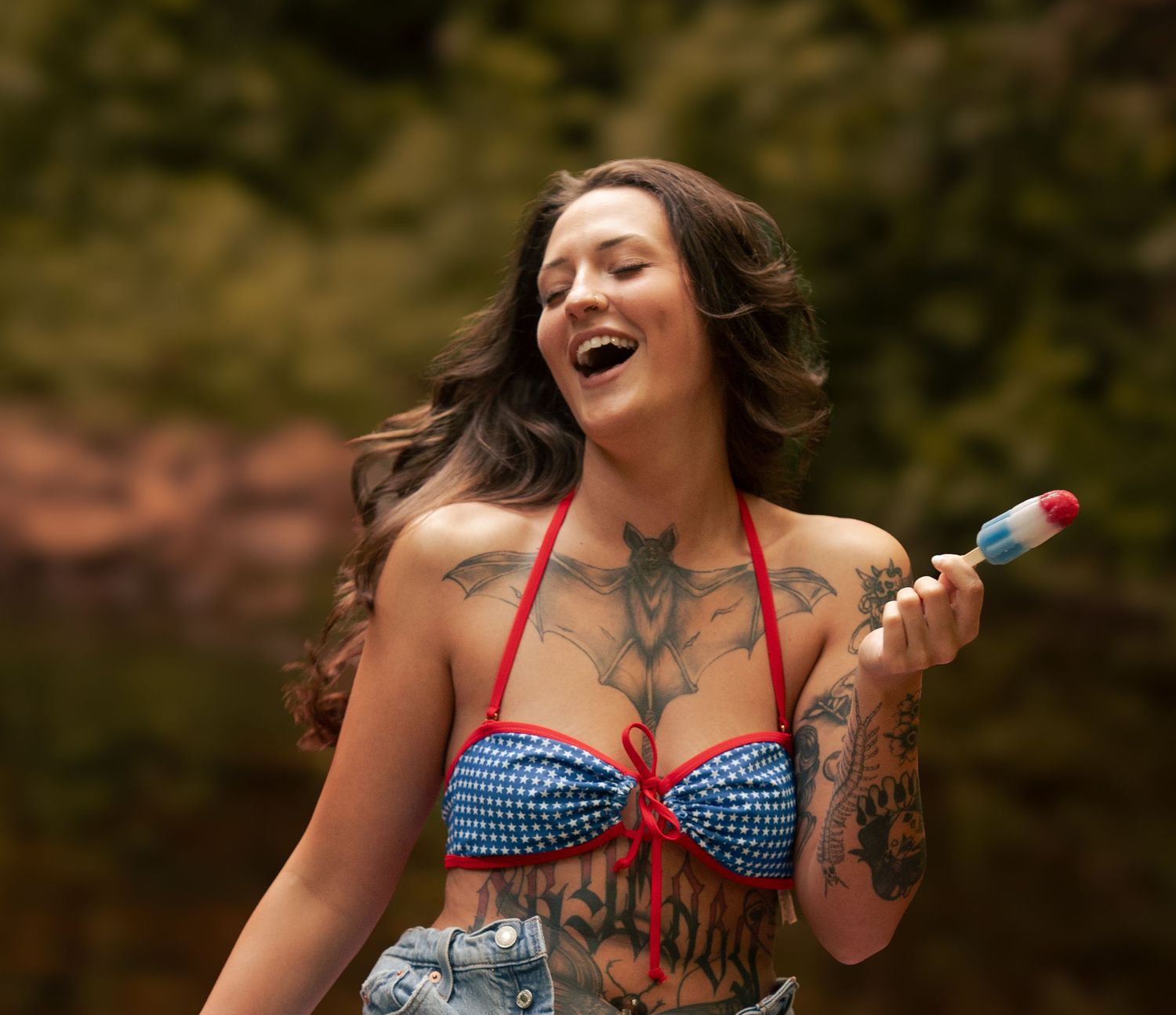 Tattooed woman in patriotic blue bikini top laughing while holding a red white and blue popsicle outdoors.