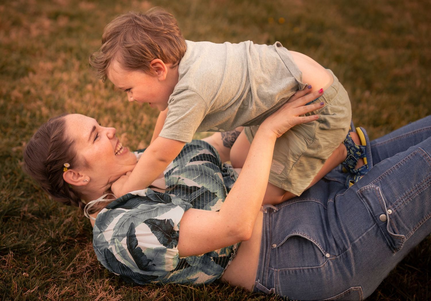 A joyful mother lying on grass playfully holding her laughing toddler son above her in a warm outdoor setting.