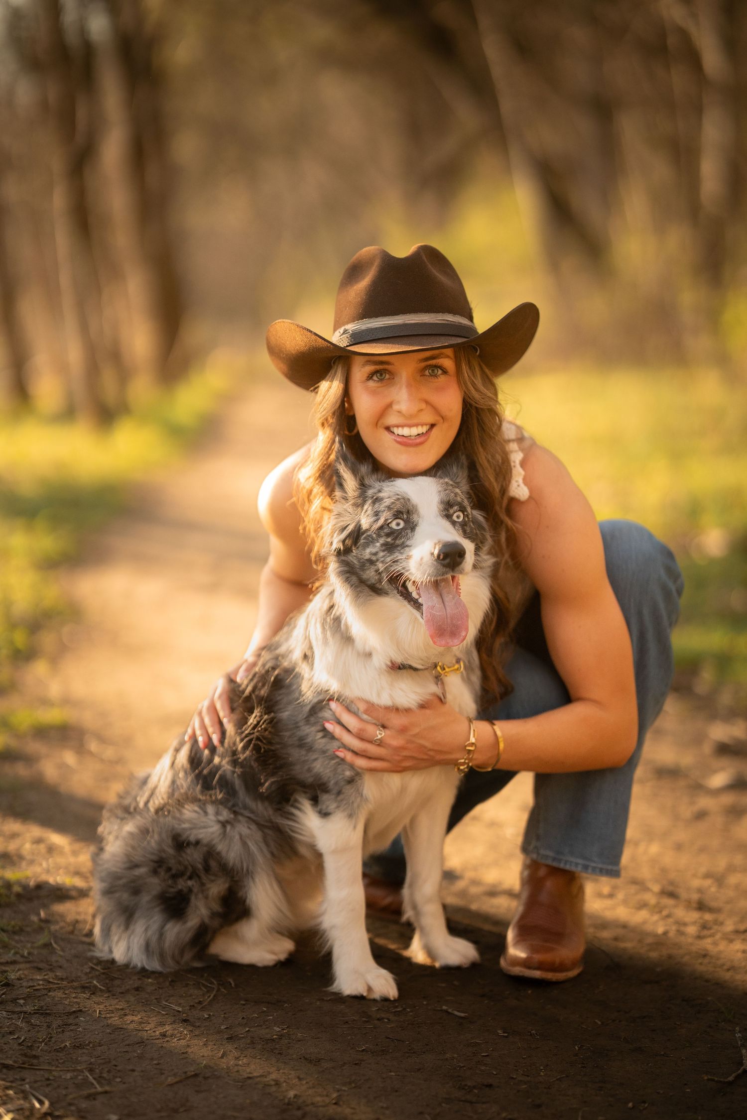 Smiling woman in cowboy hat crouching beside fluffy Australian Shepherd dog on dirt path.
