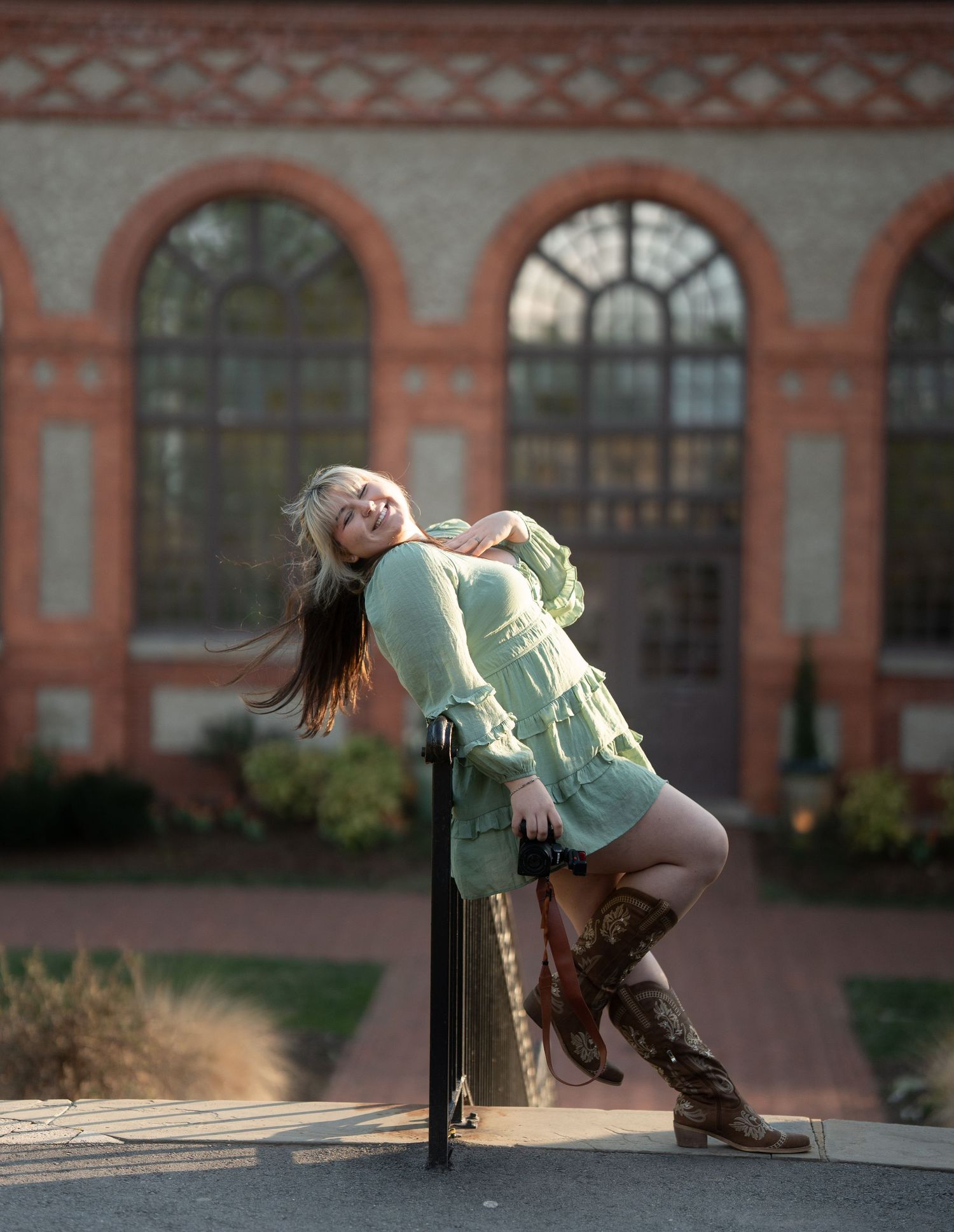 Woman in green ruffled mini dress and cowboy boots posing dramatically outside brick building.