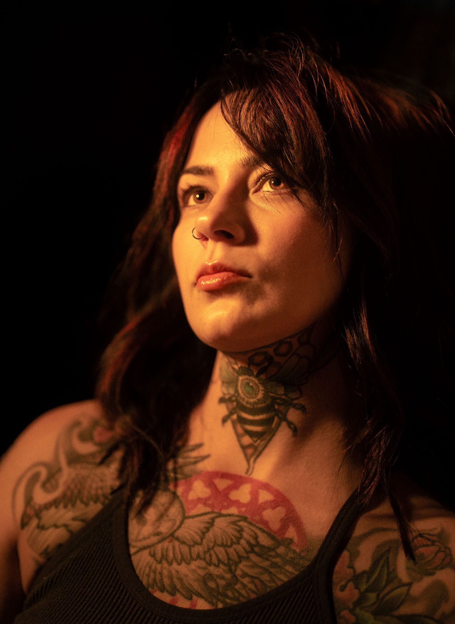 Woman with extensive neck and chest tattoos and nose ring gazes upward against dark background.