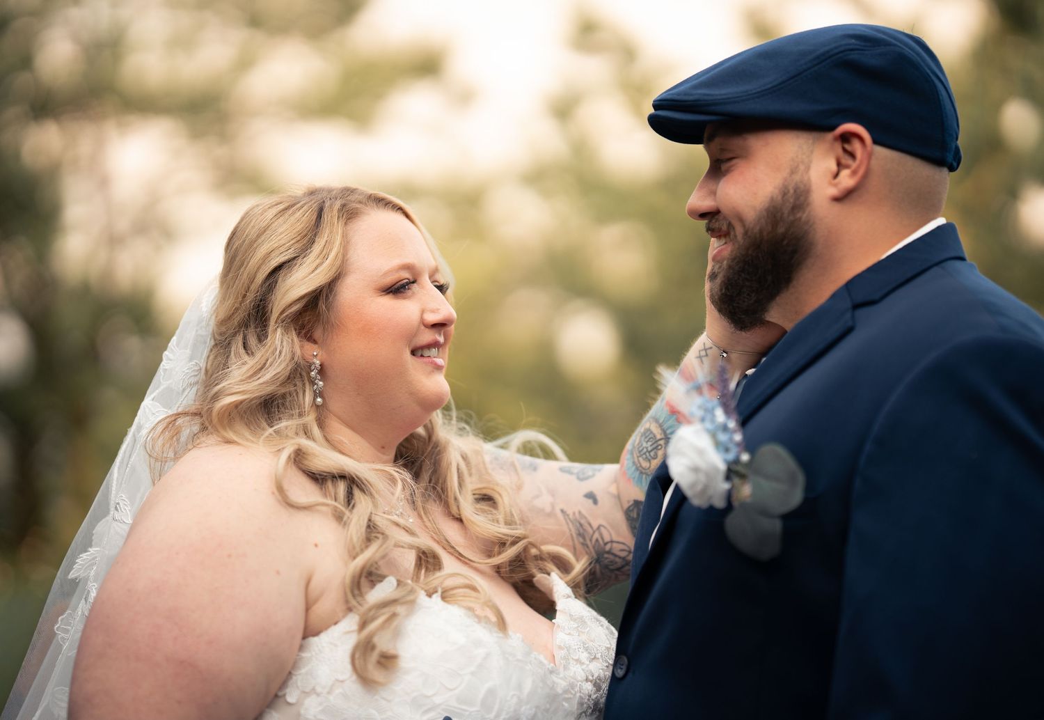 A bride and groom share a joyful moment outdoors, she in a white gown with veil, he in a navy suit with boutonniere.