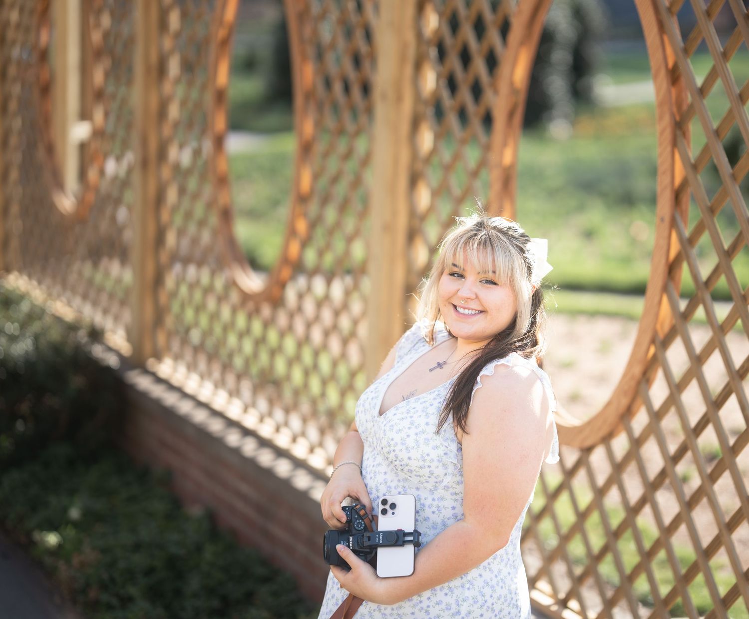 Smiling woman in floral dress holding camera in front of wooden lattice fence outdoors.