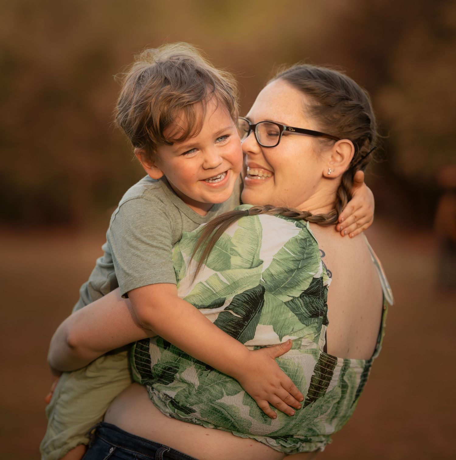 A joyful young boy hugs a smiling woman wearing glasses outdoors in warm golden light.