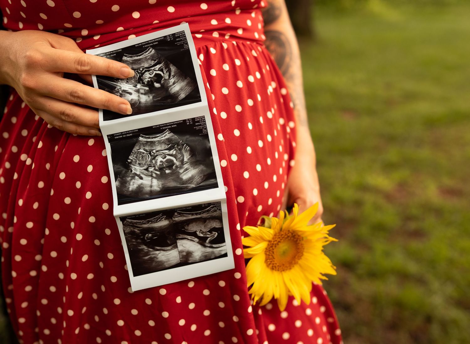 Pregnant woman in red polka dot dress holding ultrasound photos and a sunflower outdoors.