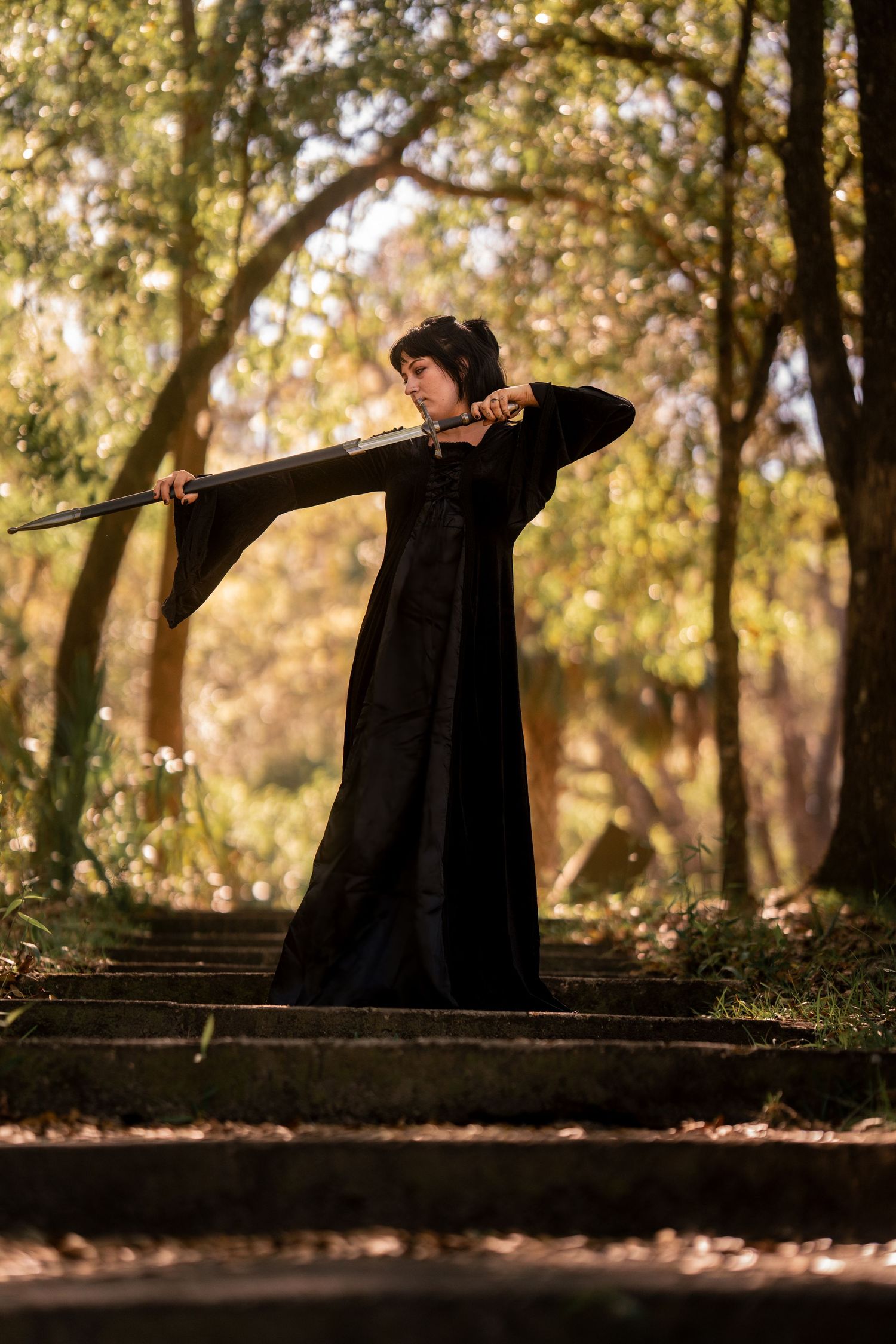 Woman in black robes wielding a long sword on stone steps in a forest setting.