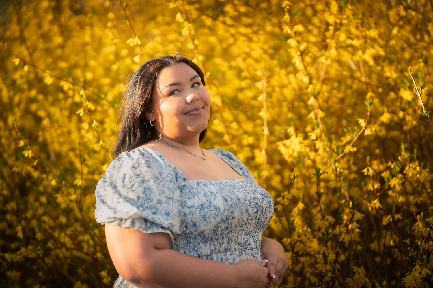 Young woman in blue floral dress smiling among vibrant yellow flowering bushes in warm sunlight.