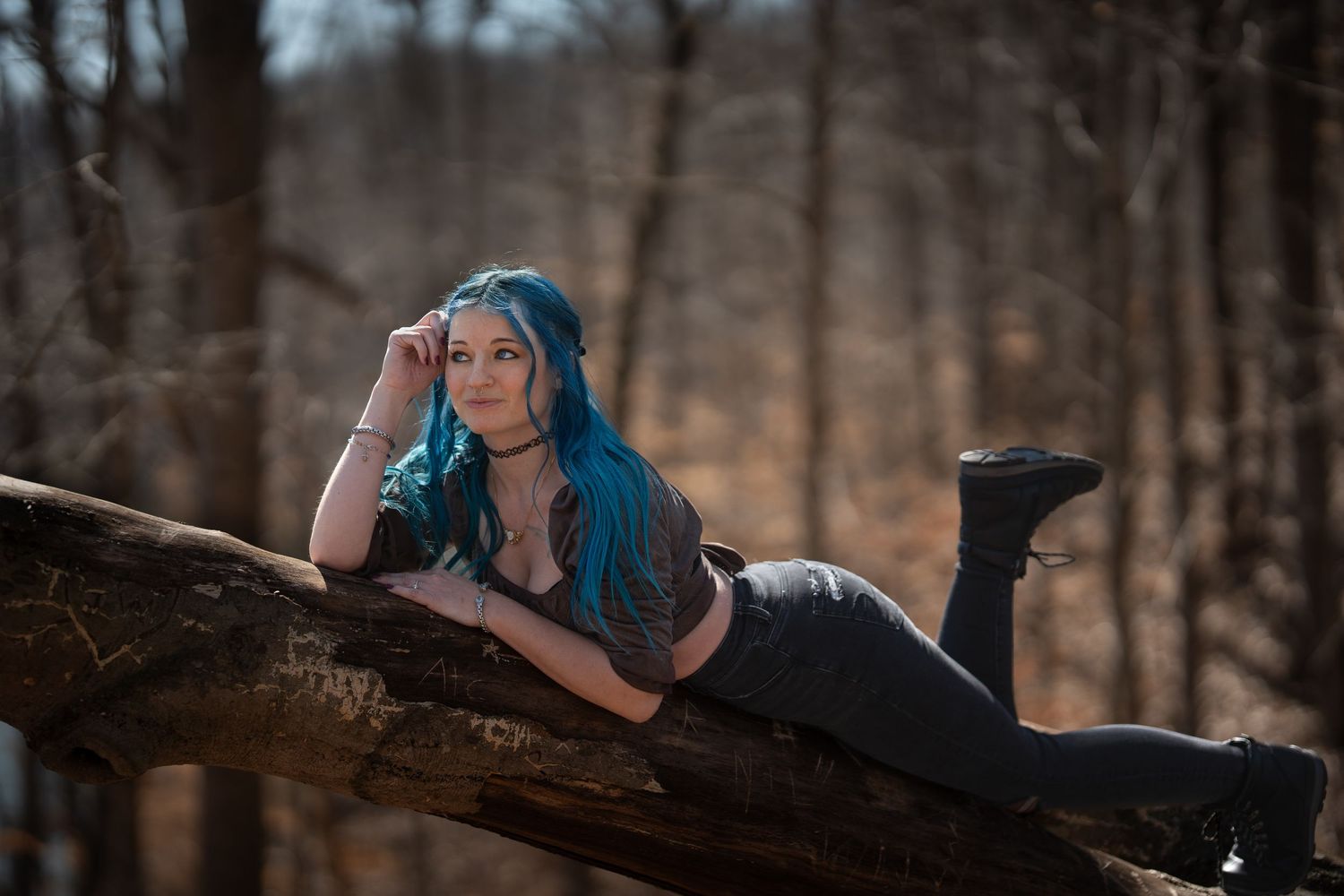Woman with blue hair reclines on fallen log in autumn forest setting.