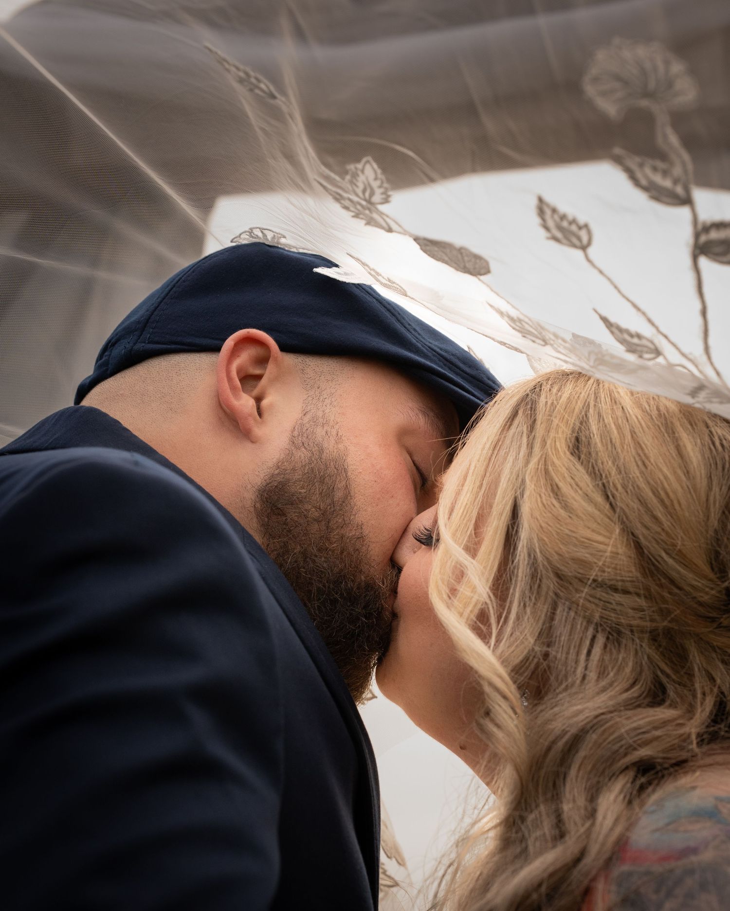 Couple sharing a romantic kiss under a sheer bridal veil, groom in navy suit and cap, bride with blonde hair.